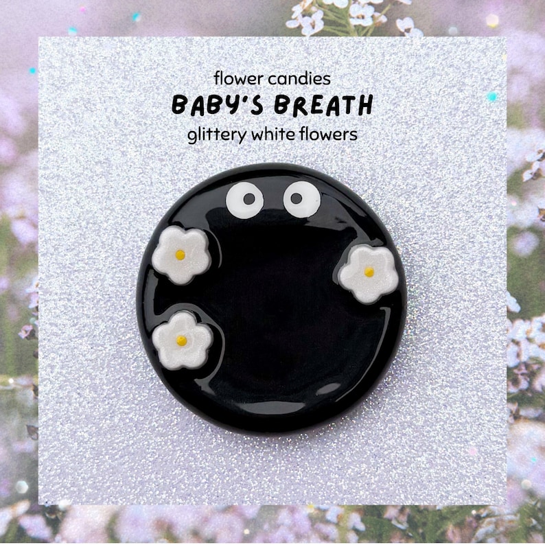 May include: A glossy black, circular decorative piece featuring white flower designs and two white eye-like shapes. The image includes the text: "flower candies BABY'S BREATH glittery white flowers."