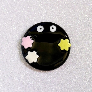 May include: A black, circular enamel pin depicting a cartoon character with two white eyes and three star-shaped accents in pink, white, and yellow. The pin has a glossy finish.