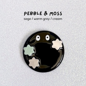 May include: A black enamel pin with a cartoon design. The pin has two white eyes and three star-shaped accents in sage, warm grey, and cream. The text "PEBBLE & MOSS" and colour names are above the pin.