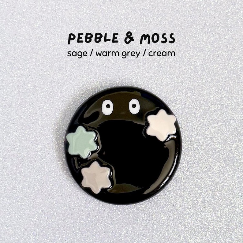 May include: A black enamel pin with a cartoon design. The pin has two white eyes and three star-shaped accents in sage, warm grey, and cream. The text "PEBBLE & MOSS" and colour names are above the pin.