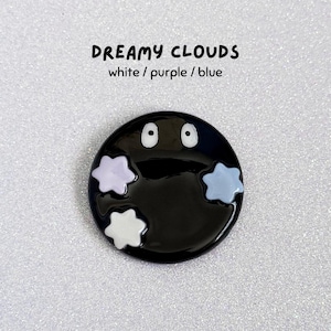 May include: A black, circular brooch featuring white eyes and three cloud-shaped details in white, purple, and blue. The text "DREAMY CLOUDS white / purple / blue" is above the brooch.