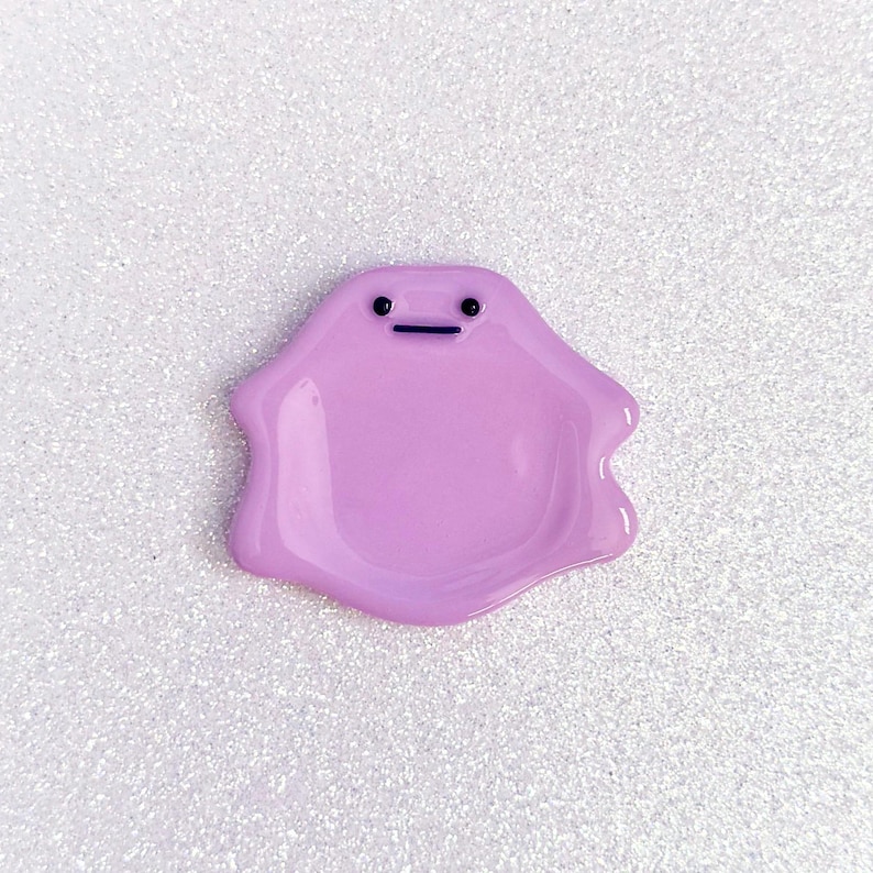 Pode incluir: Um pingente em forma de mancha de cor lavanda, com um rosto simples com dois pontos pretos para os olhos e uma linha horizontal para a boca. O pingente tem um acabamento brilhante e uma forma suave e arredondada. O fundo &eacute; uma superf&iacute;cie branca brilhante.