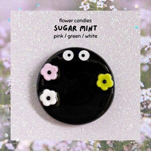 May include: A black circular pin featuring white eyes and three flower accents in pink, green, and white. The pin has the text "flower candies SUGAR MINT pink / green / white" on a white background.