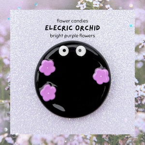 May include: A black, circular brooch featuring two white eye details and three bright purple flower motifs. The brooch has the text "flower candies ELECTRIC ORCHID bright purple flowers" on a white background.