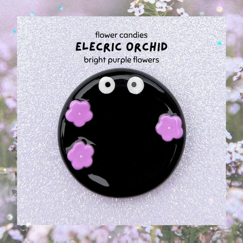 May include: A black, circular brooch featuring two white eye details and three bright purple flower motifs. The brooch has the text "flower candies ELECTRIC ORCHID bright purple flowers" on a white background.