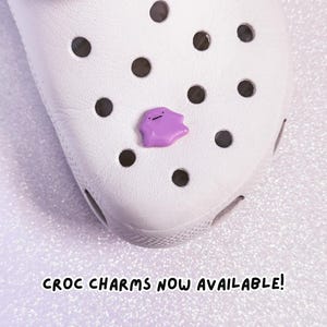 Pode incluir: Um sapato Croc branco com um charme Ditto roxo. O charme &eacute; uma pequena figura em forma de mancha com um rosto simples. O texto "CROC CHARMS NOW AVAILABLE!" est&aacute; escrito em preto na parte inferior da imagem.