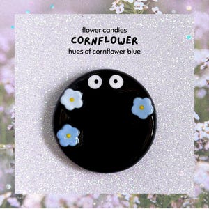 May include: A black, circular brooch featuring two white eyes and three light blue flower details with yellow centres. Text on a white background reads "flower candies CORNFLOWER hues of cornflower blue".