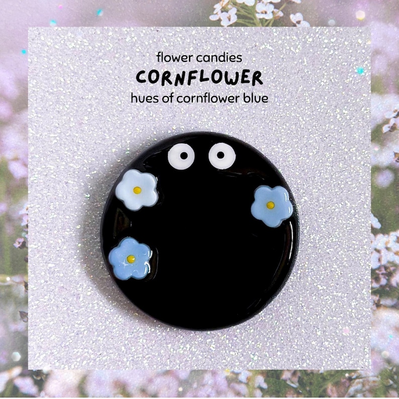May include: A black, circular brooch featuring two white eyes and three light blue flower details with yellow centres. Text on a white background reads "flower candies CORNFLOWER hues of cornflower blue".