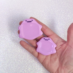 Pode incluir: Dois broches em forma de mancha de cor lavanda com tra&ccedil;os faciais pretos simples. Os broches s&atilde;o lisos e brilhantes e est&atilde;o em uma m&atilde;o aberta contra um fundo branco brilhante. Os broches t&ecirc;m um design fofo de desenho animado.
