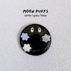 May include: A black, circular pin with the text "MOON PUFFS" above it. The pin features two white eyes and three puffy cloud shapes in white, grey, and blue. The background is a glittery, silver surface.
