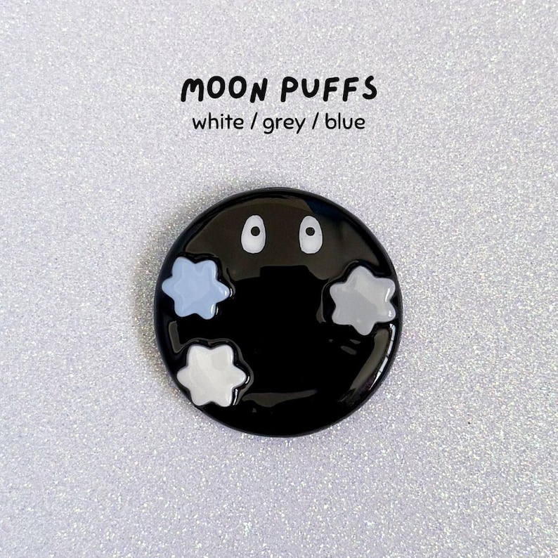 May include: A black, circular pin with the text "MOON PUFFS" above it. The pin features two white eyes and three puffy cloud shapes in white, grey, and blue. The background is a glittery, silver surface.