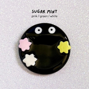 May include: A black, circular pin featuring two white eyes and three star-shaped embellishments in pink, white, and yellow. The text "SUGAR MINT" and "pink / green / white" are printed above the pin.