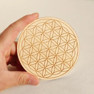 May include: A round, light-colored wooden coaster featuring a laser-etched Flower of Life pattern. The design consists of overlapping circles arranged in a symmetrical, geometric pattern. The coaster is held by a hand.