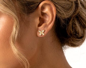 May include: A gold stud earring shaped like a butterfly, featuring four small clear stones embedded in the wings. The earring is positioned on the earlobe, showcasing a metallic gold finish and a symmetrical design with a central point.