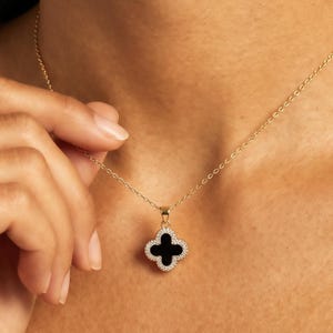 May include: A gold chain necklace featuring a four-leaf clover pendant. The pendant has a smooth black center inlay surrounded by a border of small, clear, sparkling stones. The necklace is delicate and rests against the skin with a simple clasp.