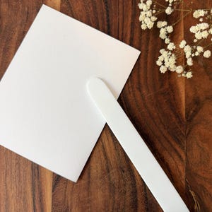 May include: A rectangular white cardstock sign attached to a long, flat white handle, creating a handheld paddle sign. The card is blank and smooth, and the handle is solid white with a rounded top edge, designed for easy holding and display.