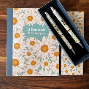 May include: A stationery set featuring a floral daisy design. The set includes a box of 12 notecards and envelopes, and a pen set with two pens. The daisy pattern is white and yellow on a light blue background.