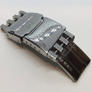 Könnte beinhalten: Ein silberner Uhrenarmbandverschluss mit der Gravur "Breitling" auf der Oberseite. Der Verschluss hat einen Druckknopf zum Öffnen und ist seitlich mit "SS MADE" gekennzeichnet.