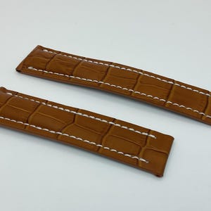 May include: Two brown watch straps with a crocodile pattern and white stitching. The straps are made of leather and are designed to attach to a watch face. The straps are a warm brown color.