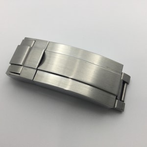 Puede incluir: Un cierre de reloj de acero inoxidable cepillado. El cierre es curvo y tiene un mecanismo plegable. El metal tiene un acabado mate y está diseñado para asegurar una correa de reloj. El cierre se muestra sobre un fondo blanco.