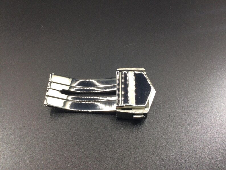 May include: A silver watch band clasp with a polished finish. The clasp has a three-piece design and a secure locking mechanism.