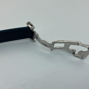 May include: Close-up of a silver watch band clasp with a black rubber strap. The clasp has a hinged design and a safety mechanism.