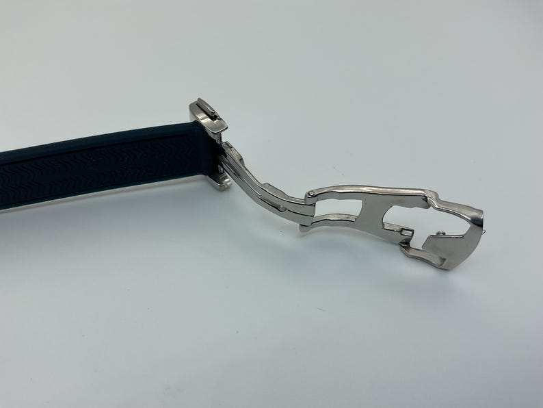 May include: Close-up of a silver watch band clasp with a black rubber strap. The clasp has a hinged design and a safety mechanism.