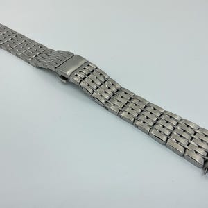 Puede incluir: Una correa de reloj de metal plateado con cierre. La correa está hecha de pequeñas piezas de metal unidas.