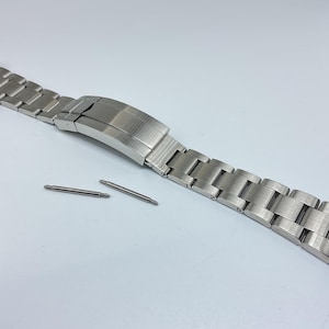 Puede incluir: Una correa de reloj plateada con acabado cepillado y cierre. La correa está colocada plana sobre una superficie blanca con dos pasadores.