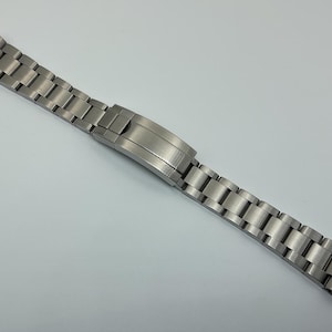 Puede incluir: Una correa de reloj plateada con acabado cepillado y un eslabón central pulido.