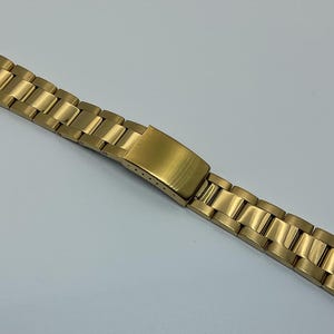 Puede incluir: Una correa de reloj dorada con un acabado cepillado. La correa está compuesta por eslabones rectangulares conectados por eslabones más pequeños y articulados. El cierre es de tipo desplegable, también en dorado. La correa está diseñada para fijarse a una caja de reloj.