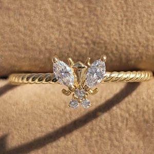 May include: A gold-colored ring featuring a butterfly design. The butterfly has two marquise-cut stones for wings and smaller round stones. The band has a twisted rope design.