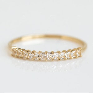 Pode incluir: Um delicado anel de ouro com uma fileira de pedras preciosas claras, de corte quadrado. O anel tem uma banda fina e as pedras são uniformemente espaçadas, criando um brilho contínuo. O anel está sobre um fundo branco suave.