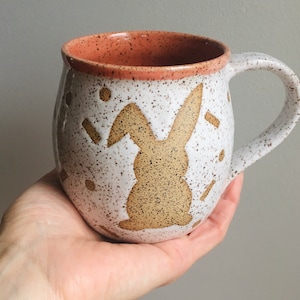 May include: A speckled white ceramic mug with a brown bunny silhouette and accents. The interior and rim are a warm orange-brown color. The mug has a large handle and is held in a hand.