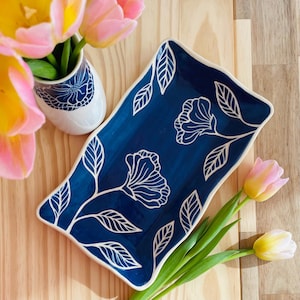 May include: A rectangular, navy blue ceramic serving platter with white floral and leaf designs. The platter has a wavy edge and is next to a small white vase with a blue floral pattern and pink and yellow tulips.