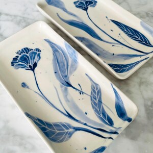 May include: Two rectangular ceramic serving platters with a white background. Each platter features a blue floral design with leaves and a flower. The design is hand-painted with varying shades of blue, creating a watercolor effect.