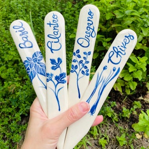 May include: Four ceramic plant markers with blue and white designs. The markers are labeled "Basil", "Cilantro", "Oregano", and "Chives".