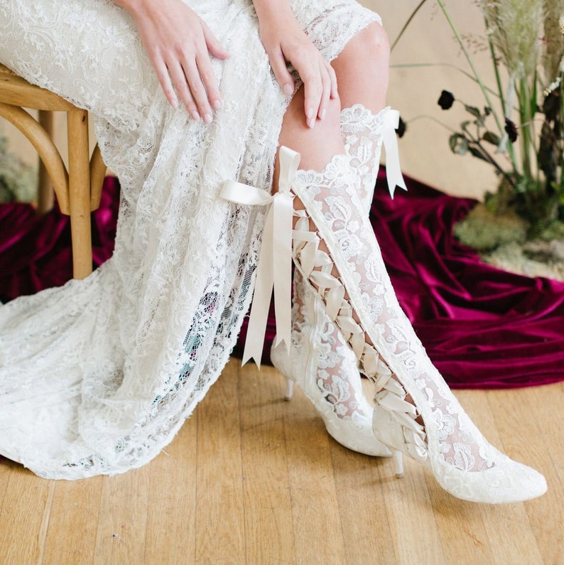 Puede incluir: Botas de novia de encaje blanco con un lazo de cinta de sat&eacute;n. Las botas tienen un delicado patr&oacute;n floral y un tac&oacute;n alto.