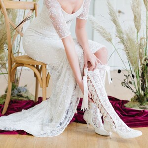 Puede incluir: Una novia con un vestido de novia de encaje blanco y botas de encaje blanco con lazos de cinta. Est&aacute; sentada en una silla de madera con una tela de terciopelo rojo en el suelo.
