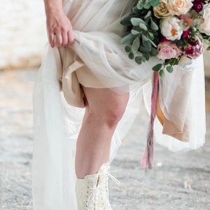 Pode incluir: Uma noiva usando um vestido de noiva de renda branca e botas brancas com cadar&ccedil;os. Ela est&aacute; segurando um buqu&ecirc; de flores rosa, brancas e verdes.