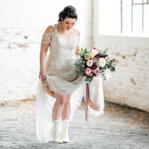 Pode incluir: Uma noiva usando um vestido de noiva de renda branca com um buqu&ecirc; de flores rosa, brancas e bord&ocirc;. Ela est&aacute; usando botas brancas e est&aacute; segurando o buqu&ecirc; na frente dela.