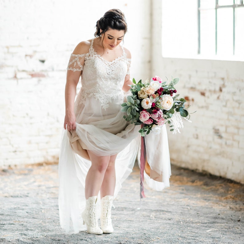 Pode incluir: Uma noiva usando um vestido de noiva de renda branca com um buqu&ecirc; de flores rosa, brancas e bord&ocirc;. Ela est&aacute; usando botas brancas e est&aacute; segurando o buqu&ecirc; na frente dela.