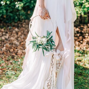 Puede incluir: Una novia con un vestido de novia blanco con una bota de encaje y sosteniendo una corona de flores. La corona est&aacute; hecha de flores blancas y vegetaci&oacute;n.