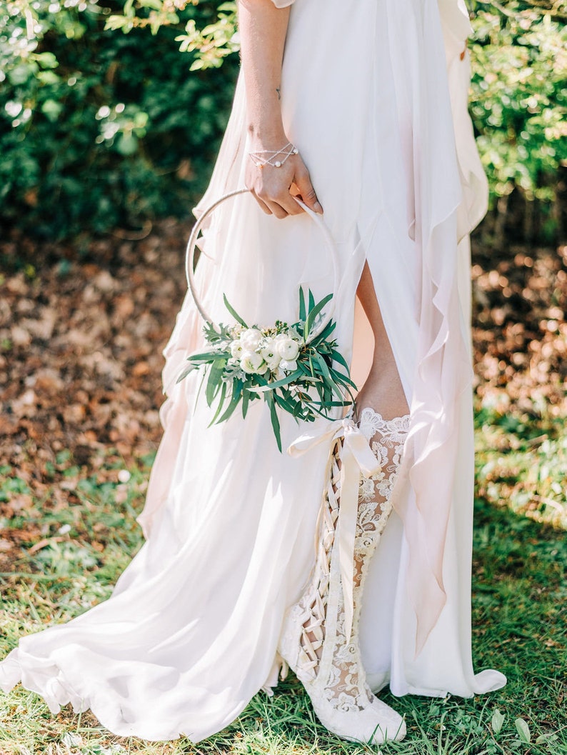 Puede incluir: Una novia con un vestido de novia blanco con una bota de encaje y sosteniendo una corona de flores. La corona est&aacute; hecha de flores blancas y vegetaci&oacute;n.