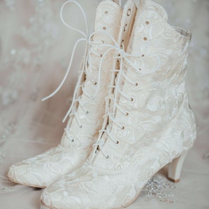 Pode incluir: Um par de botas de tornozelo brancas de renda com um delicado padr&atilde;o floral. As botas t&ecirc;m um salto alto e fechos de renda.