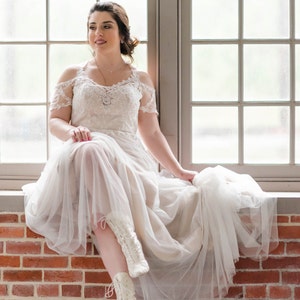 Pode incluir: Uma noiva vestindo um vestido de noiva branco de renda com uma saia de tule e botas brancas de renda. Ela est&aacute; sentada em uma parede de tijolos na frente de uma janela.
