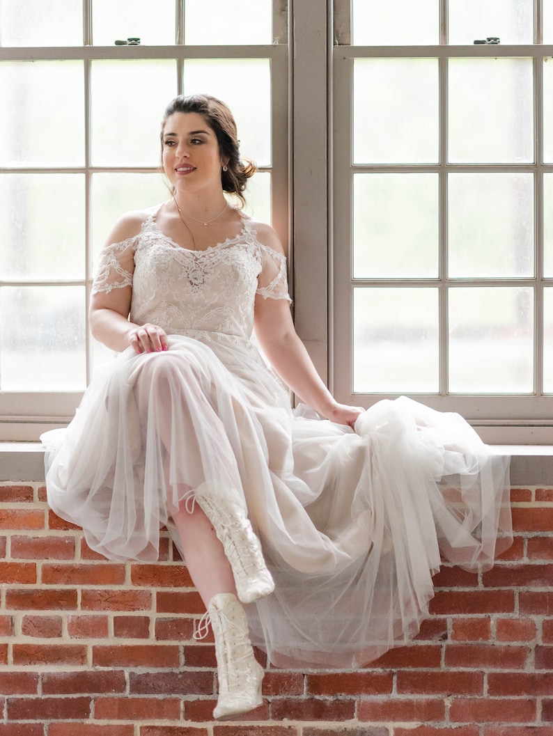 Pode incluir: Uma noiva vestindo um vestido de noiva branco de renda com uma saia de tule e botas brancas de renda. Ela est&aacute; sentada em uma parede de tijolos na frente de uma janela.