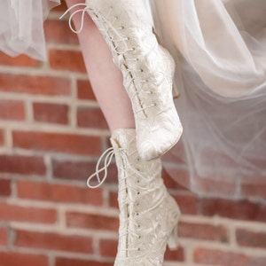 Pode incluir: Um par de botas brancas de renda com salto alto. As botas s&atilde;o feitas de um tecido delicado com um padr&atilde;o floral. As botas s&atilde;o usadas por uma pessoa em p&eacute; em uma superf&iacute;cie de tijolo.