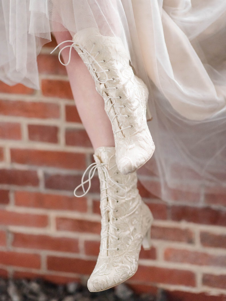 Pode incluir: Um par de botas brancas de renda com salto alto. As botas s&atilde;o feitas de um tecido delicado com um padr&atilde;o floral. As botas s&atilde;o usadas por uma pessoa em p&eacute; em uma superf&iacute;cie de tijolo.