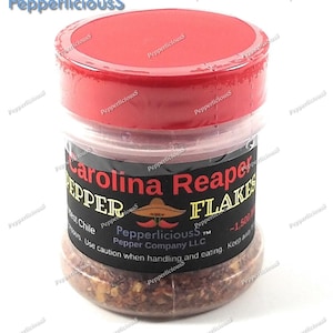 May include: A clear glass jar with a red lid filled with Carolina Reaper pepper flakes. The jar has a black label with white text that reads "Carolina Reaper Pepper Flakes" and "Pepperlicious Pepper Company LLC". The label also includes a small image of a chili pepper wearing a sombrero.